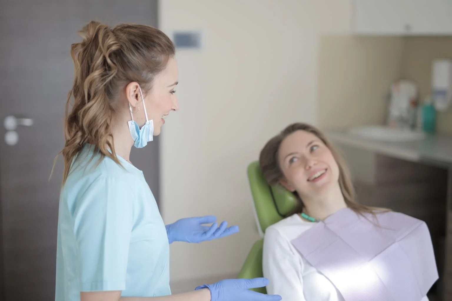 A dentist in light blue scrubs talking with a smiling patient in a dental chair