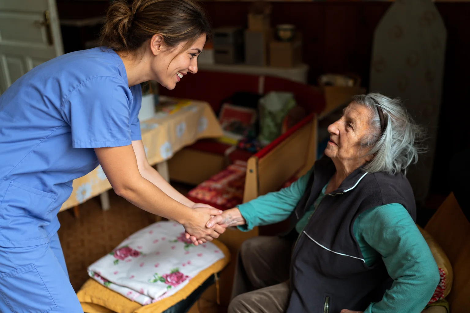 A younger caregiver in blue scrubs holding an older woman’s hand in a home setting